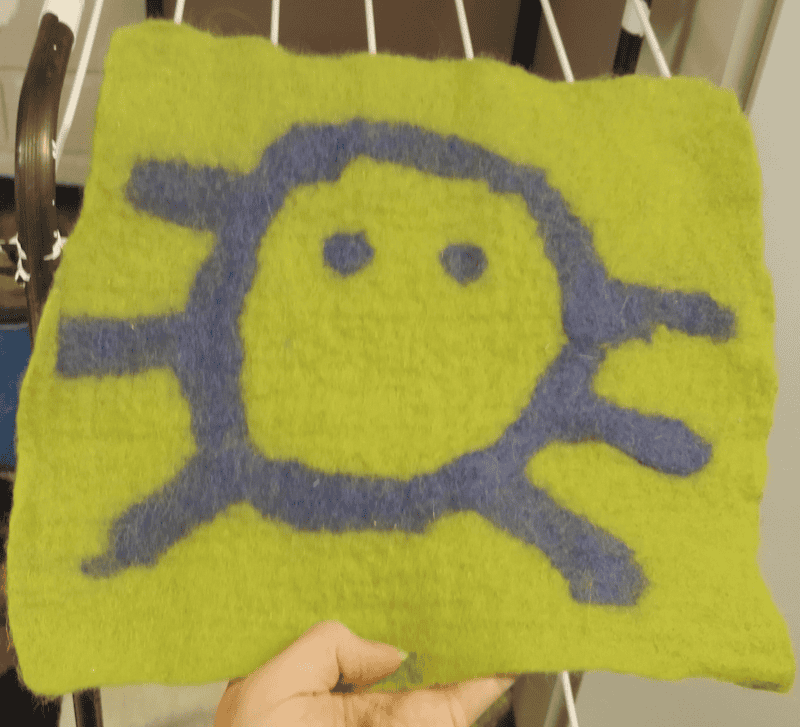 Felt image of a 6 legged purple spider on a green background.